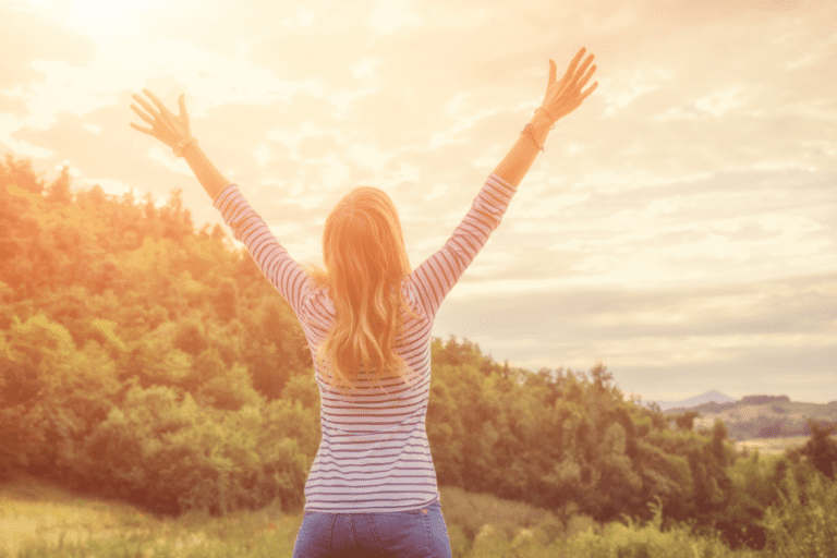 A woman from behind with her arms raised high toward a bright, sunlit sky over a forest.