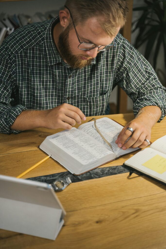 A bearded man with glasses sitting at a wooden table, reading a Bible and taking notes.