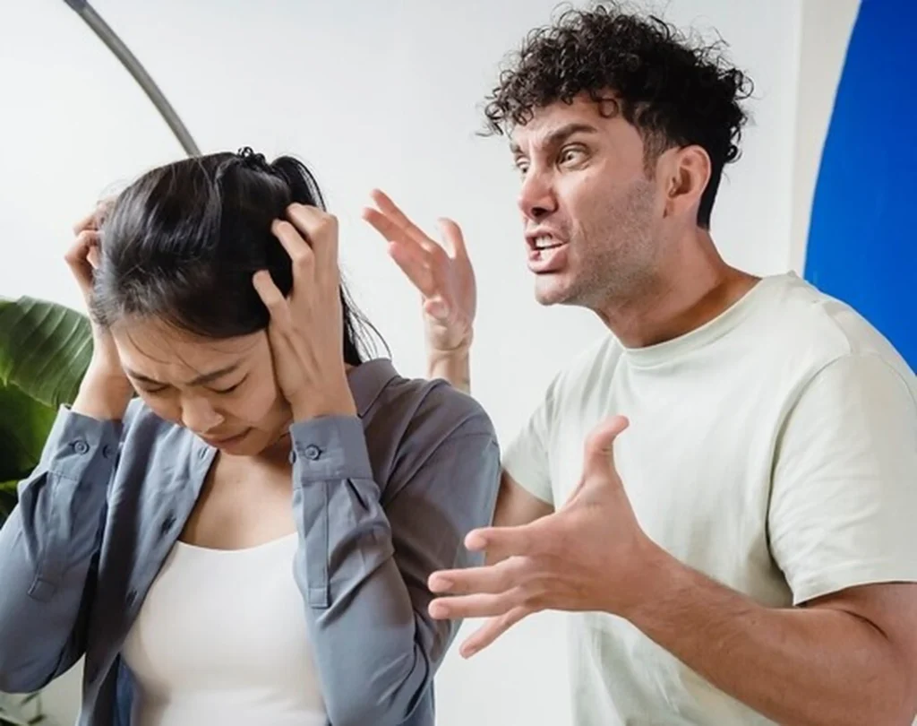 A man shouting aggressively at a woman who is covering her ears and cowering in distress.