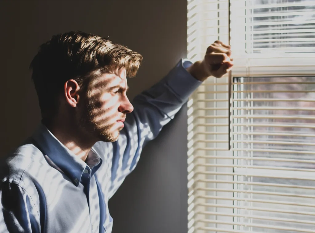 A man in a blue shirt leaning against a window, his face partially covered by the shadows of horizontal blinds.