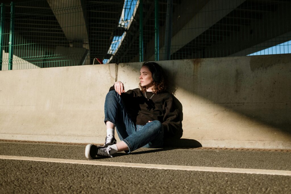 A young woman with headphones sitting on the ground, leaning against a concrete barrier under a highway overpass.